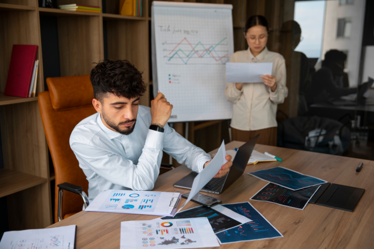 A man reviews data charts at a desk while a woman stands behind him, studying documents in a modern office.